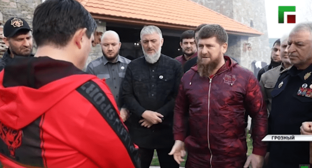 Асланбек Ахметханов (в красном) стоит перед Рамзаном Кадыровым. Стоп-кадр сюжета ЧГТРК "Грозный" "Глава ЧР жестко отчитал Асламбека Ахметханова", https://youtu.be/IUHeFy2cpfk